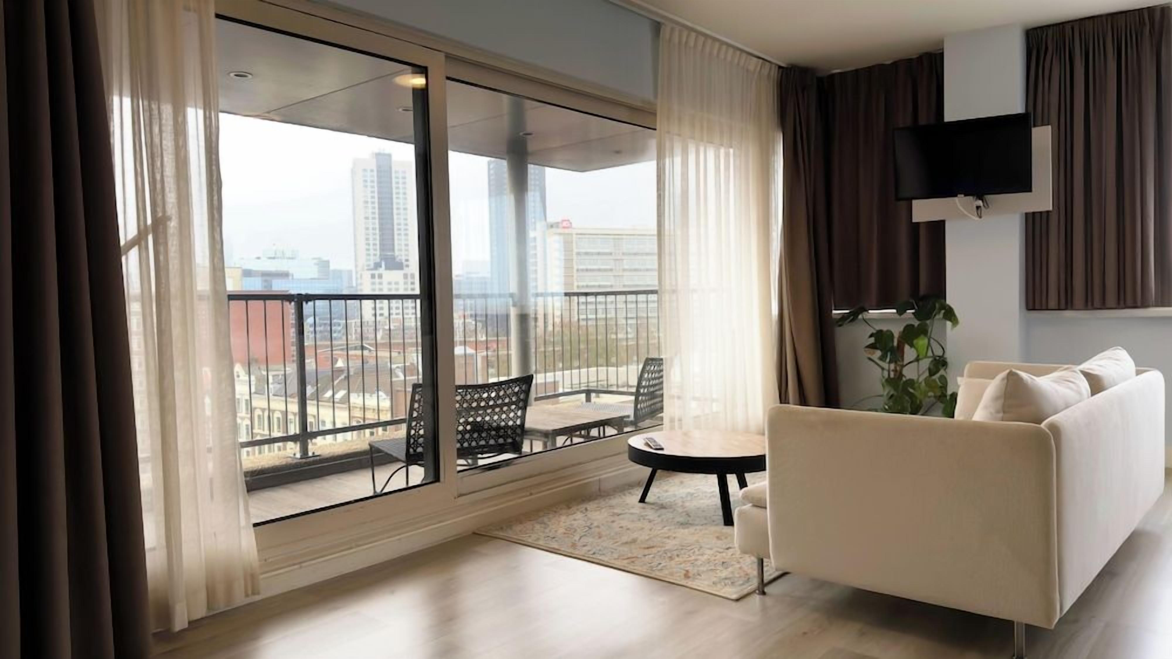 A modern living room with a sofa, a small table, and a chair, a balcony with a view of the city, and a flat-screen TV mounted on the wall.