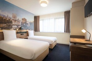 Et hotelværelse med to senge, et bybillede på væggen, et skrivebord med lampe samt et TV.