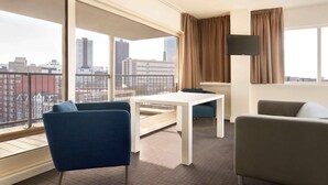 A modern hotel room with a large window, a white table, two blue chairs, a beige sofa, and a flat-screen TV. - Rotterdam Teleport Hotel (Rotterdam)