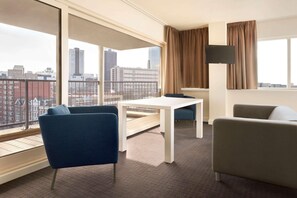 Habitación de hotel moderna con un ventanal grande, una mesa blanca, dos sillas azules, un sofá beige y un televisor de pantalla plana.