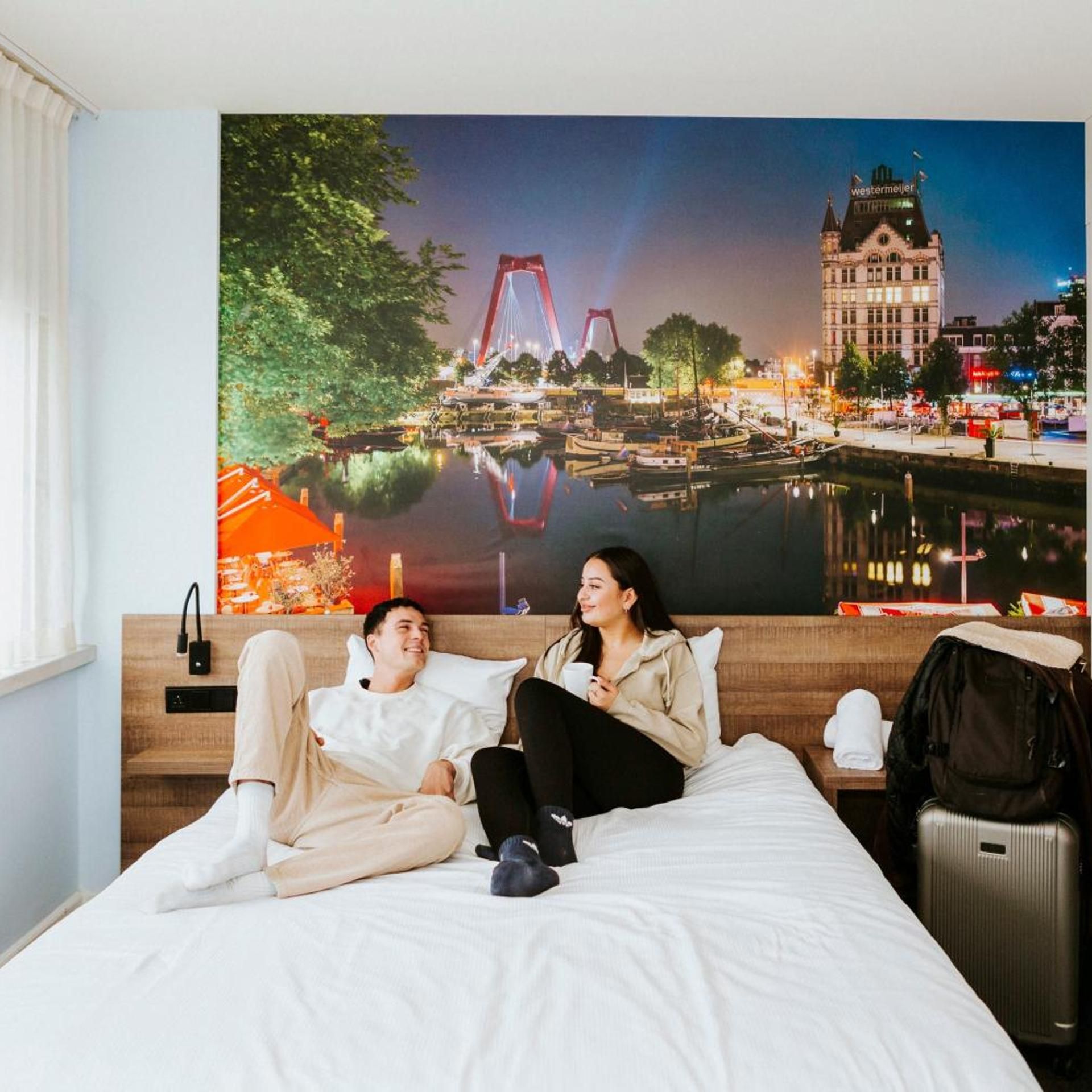 A hotel room with a large bed, a nightstand, a wall-mounted lamp, and a poster of a cityscape.