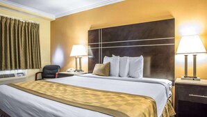 Een hotelkamer met een groot bed, twee bedlampen, een hoofdbord, een bureau met lamp, een stoel en een raam met gordijnen.