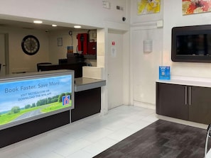A hotel reception area with a digital screen displaying a booking message, a clock on the wall, and a fire extinguisher. - Motel6 Hagerstown, MD (Hagerstown)