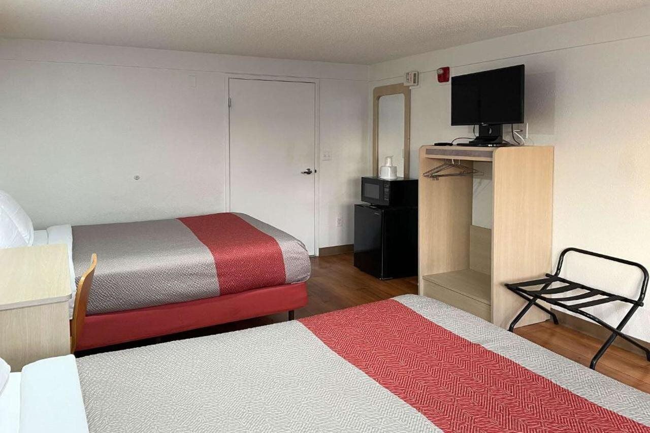 Quarto de hotel com cama, escrivaninha, cadeira, televisão e guarda-roupa.
