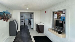 A hallway with a black and white tiled floor, a trash bin, a vending machine, and a television mounted on the wall. - Motel6 Hagerstown, MD (Hagerstown)