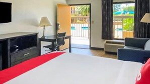 Une chambre d’hôtel équipée d’un lit, d’un bureau, d’une chaise, d’une télévision et d’une fenêtre donnant sur une piscine et un espace verdoyant.