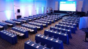 Une salle de conférence avec des rangées de tables, chacune étant dotée de plaques nominatives et de bouteilles d’eau, ainsi qu’une scène équipée d’un écran de présentation.