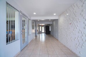 Een lange, smalle gang met betegelde vloer, een brandblusser aan de muur en een deur aan het einde die naar een andere kamer leidt.