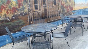 Eine Terrassengaststätte mit Metalltischen und -stühlen, einem Wandbild von Haus und Bäumen und Blick auf ein Gewässer.