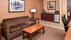 Habitación de hotel con sofá, una mesita de centro de madera, una mesita auxiliar con lámpara, una kitchenette con microondas y un cuadro en la pared.