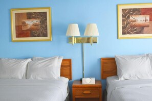 Ein Hotelzimmer mit zwei Betten, einem Nachttisch mit Lampe und zwei gerahmten Kunstwerken an der Wand.