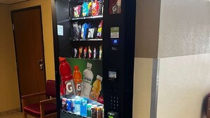 A vending machine with various beverages and snacks, a wooden chair, and a door in a room with tiled flooring. - Surestay by Best Western Glendive Yellowstone River (Glendive)