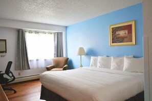 Habitación de hotel con una cama grande, un escritorio con silla, una ventana con cortinas, una lámpara y un cuadro en la pared.