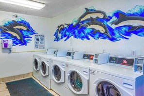 Una sala de lavandería con varias lavadoras y secadoras, un mural en la pared con delfines y un cartel que indica lavado sencillo.