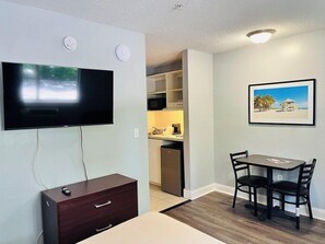 Una habitación con un televisor montado en la pared, una pequeña mesa con sillas, un armario de madera y un cuadro enmarcado.