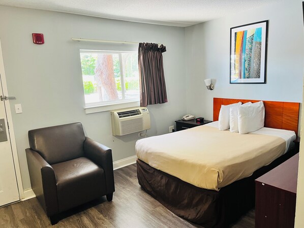 Habitación de hotel con cama, una silla, mesita de noche con teléfono y lámpara, ventana con cortinas y aire acondicionado instalado en la pared.