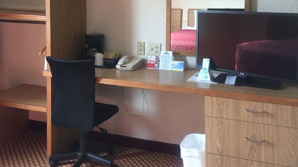 A hotel room with a wooden desk, a black office chair, a bed with a red blanket, a small trash bin, and a wall-mounted mirror. - Motel 6 Coraopolis, PA Pittsburgh Airport (Coraopolis)
