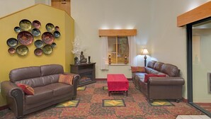 A living room with two brown leather sofas, a coffee table, a wall-mounted decorative plate collection, a small statue, and a window with blinds. - Motel 6 Coraopolis, PA Pittsburgh Airport (Coraopolis)