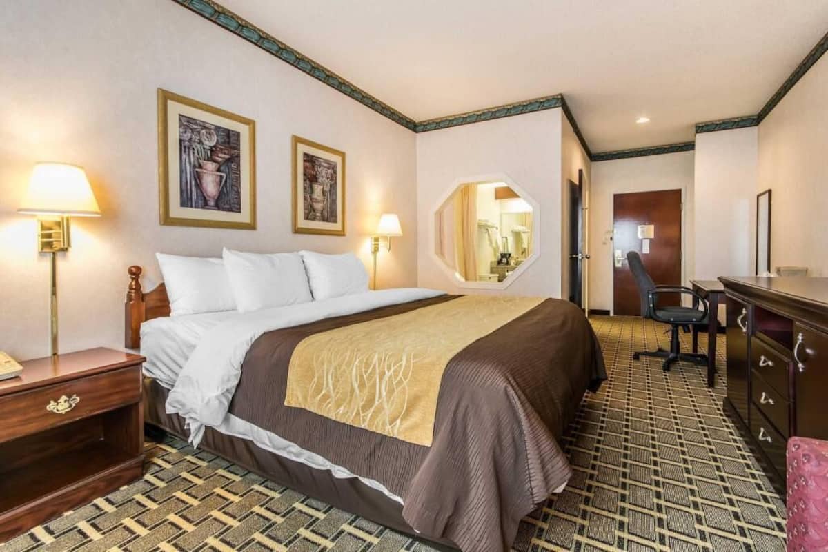 A hotel room with a bed, bedside lamps, a desk, a chair, a dresser, and a mirror.