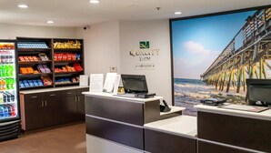 A hotel reception area with a large poster of a pier, a refrigerator stocked with snacks, and a counter with a computer. - Quality Inn Whiteville North (Whiteville)