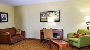 A hotel room with a brown patterned carpet, a brown sofa, a green armchair, a wooden desk with a chair, a lamp, and a framed picture of a tree. - Quality Inn Whiteville North (Whiteville)