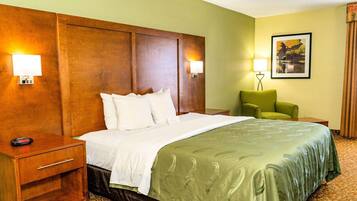 A hotel room with a large bed, a wooden headboard, two bedside lamps, a green armchair, and a framed picture on the wall.
