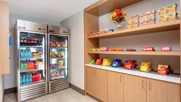 A kitchenette with two refrigerators stocked with beverages and snacks, and shelves above holding packaged food items.