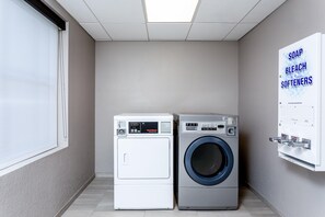 A laundry room with two washing machines, a window, and a wall-mounted soap and bleach dispenser. - Holiday Inn Express & Suites Palm Coast - I-95 (Palm Coast)