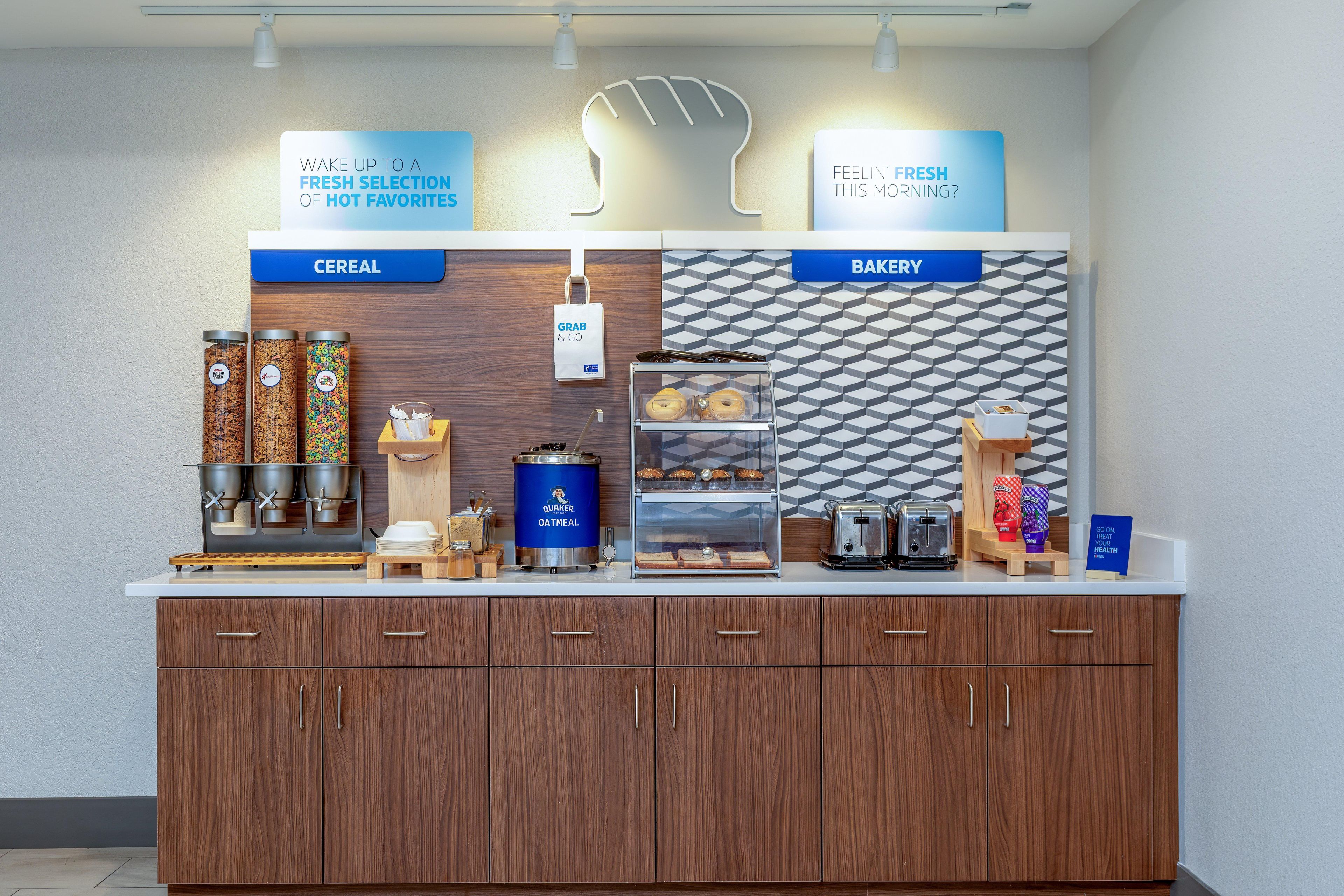 Ein Frühstücksbuffet mit beschrifteten Bereichen für Müsli und Backwaren, inklusive verschiedener Speisen und einem Schild mit der Aufschrift 'zum Mitnehmen'.