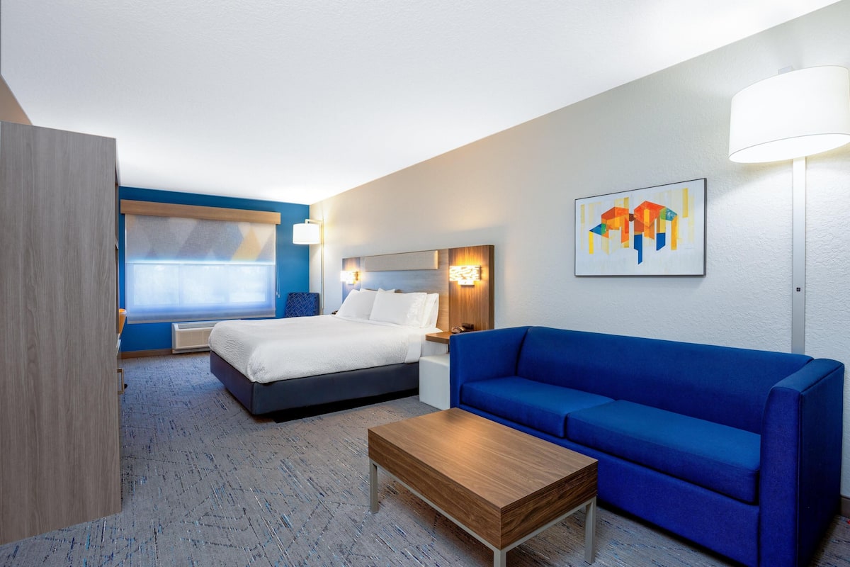 A hotel room with a bed, a blue sofa, a wooden coffee table, a wall-mounted lamp, and a painting.