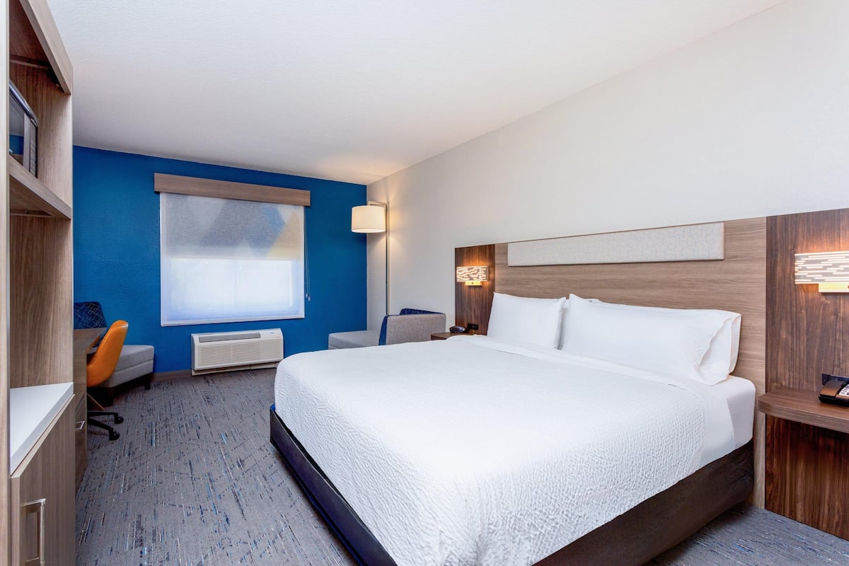 A hotel room with a large bed, a desk with a chair, a window with blinds, a wall-mounted lamp, and a bedside table with a phone.
