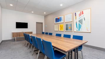 Un salón de conferencias con una larga mesa, sillas azules, un televisor de pantalla plana y obras de arte enmarcadas en la pared.