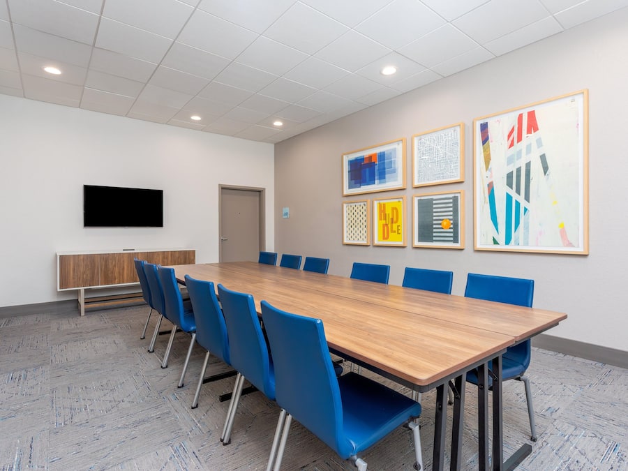 A conference room with a long table, blue chairs, a flat-screen TV, and framed artwork on the wall.