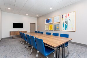 Un salón de conferencias con una larga mesa, sillas azules, un televisor de pantalla plana y obras de arte enmarcadas en la pared.