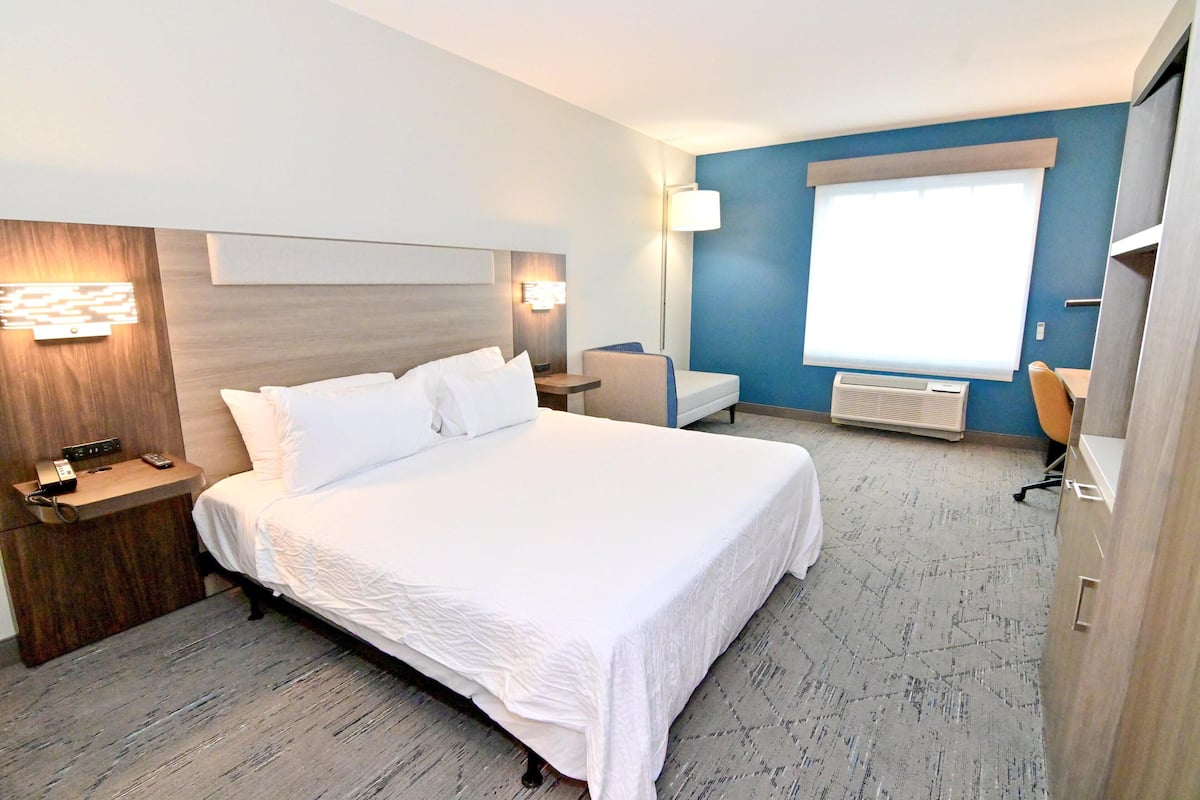 A hotel room with a large bed, bedside tables, a chair, a desk, a lamp, and a window.
