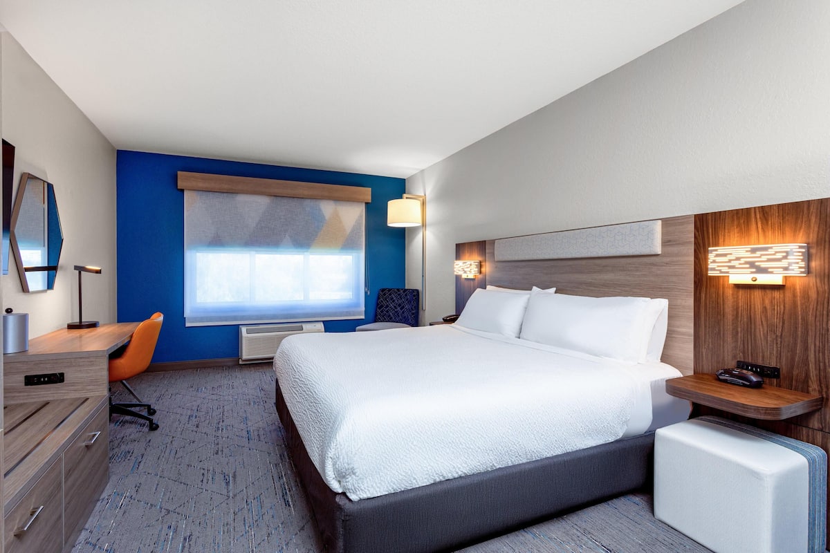 A hotel room with a large bed, a desk, a chair, a dresser, a window, and a wall-mounted lamp.