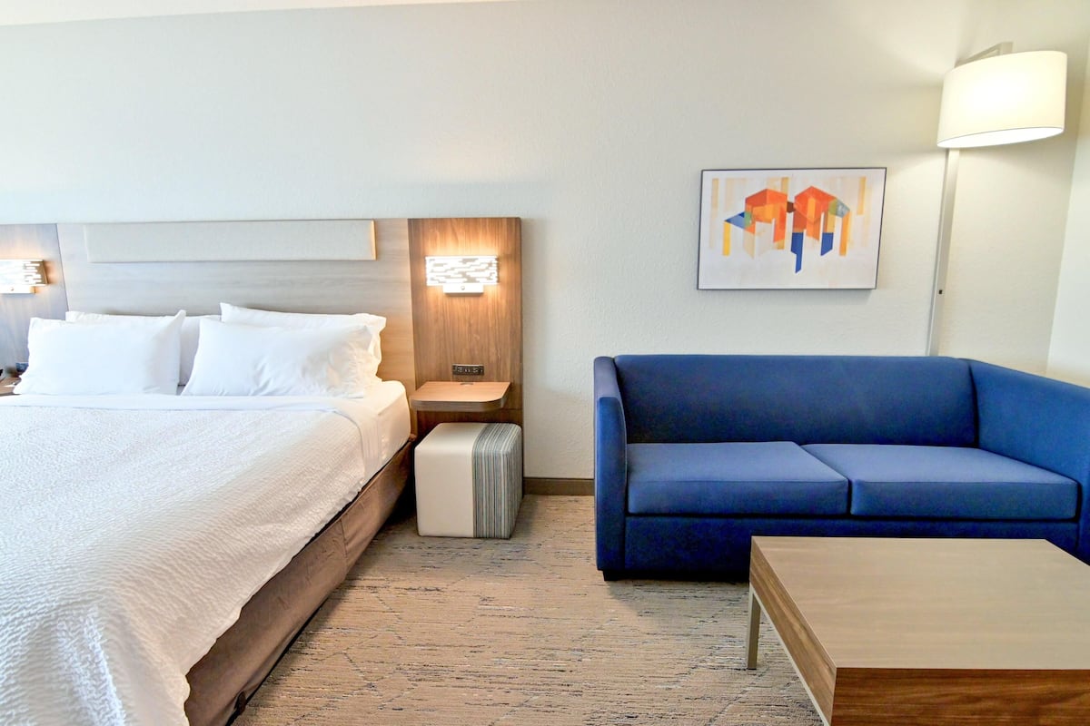 A hotel room with a bed, a blue sofa, a wooden coffee table, a nightstand with a lamp, and a wall-mounted artwork.