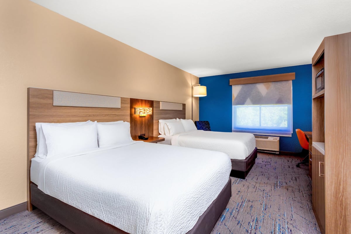 A hotel room with two beds, a desk, a chair, a window with blinds, and a wall-mounted lamp.