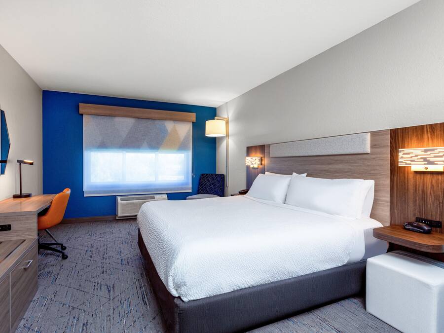 Une chambre d’hôtel avec un grand lit, un bureau, une chaise, une commode, une fenêtre et une lampe fixée au mur.