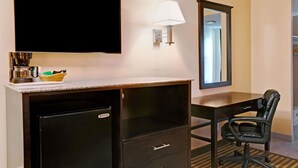 Ein Hotelzimmer mit Flachbildfernseher, einer Kaffeemaschine, einem Schreibtisch mit Schubladen, einem Sessel und einem Spiegel.