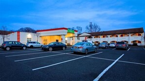 Ein Parkplatz mit mehreren parkenden Autos vor einem Gebäude mit rotem und weißem Putz, einem grünen Schild und einem mehrstöckigen Bauwerk mit orangenen Akzenten.