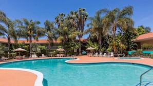 A swimming pool with lounge chairs and umbrellas, surrounded by palm trees and a building with a red-tiled roof. - Uptown Oasis San Jose Airport (San Jose)
