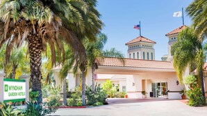 A hotel entrance with a sign, palm trees, and a building with a tiled roof. - Uptown Oasis San Jose Airport (San Jose)