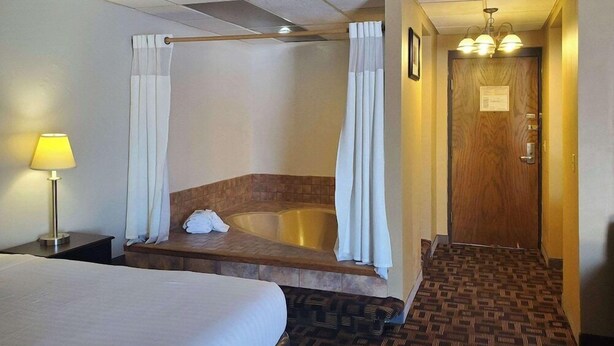 Quarto de hotel com uma cama grande, um banco de madeira, uma banheira e um corredor que leva a uma porta.