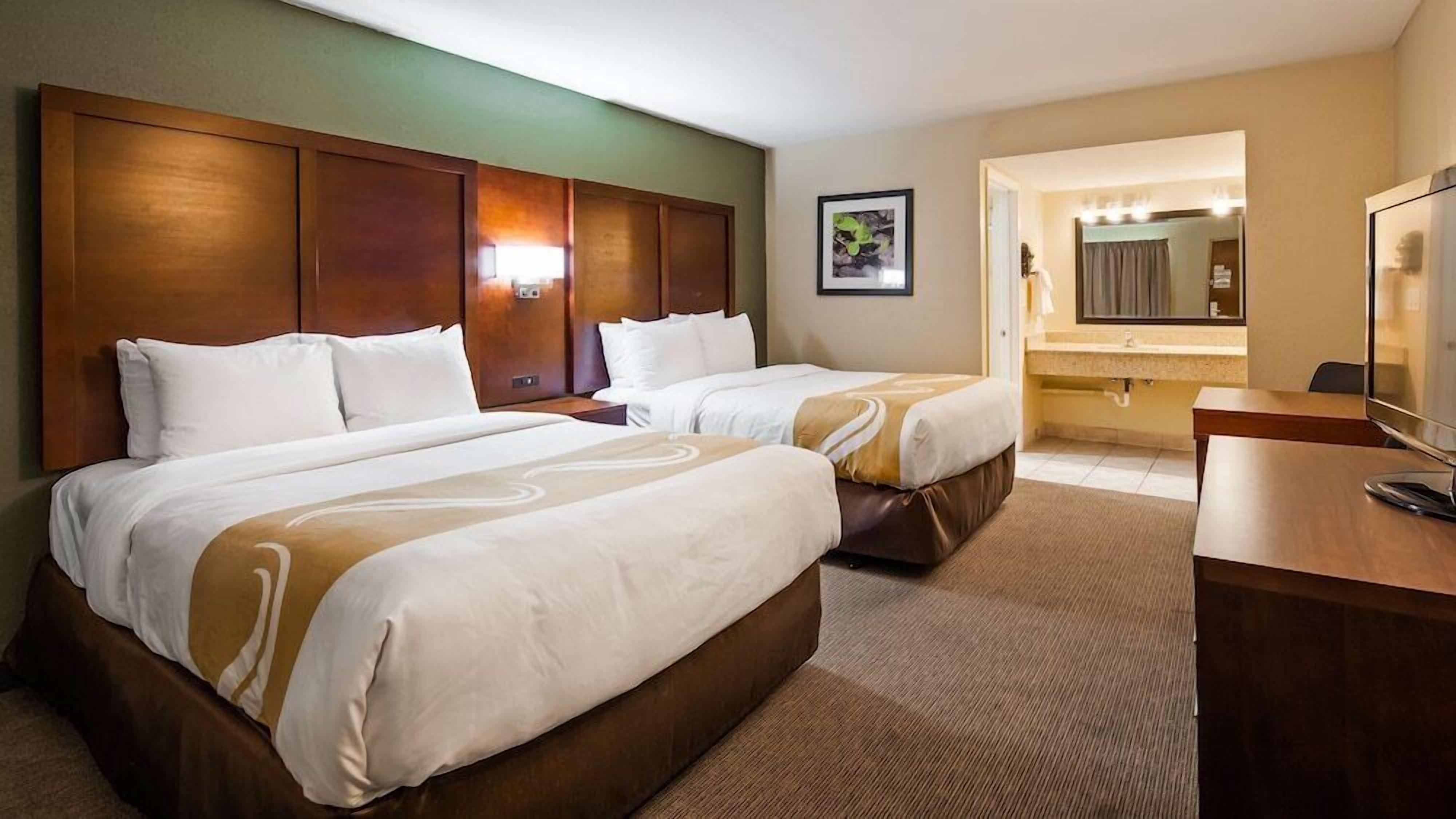 Habitación de hotel con dos camas, un escritorio, un espejo y un baño con lavamanos y espejo.