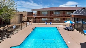 A rectangular swimming pool with a clear blue water, surrounded by a metal fence. There are two lounge chairs and a table with a blue umbrella. A multi-story building with balconies is visible in the background. - Motel6 Willcox, AZ (Willcox)