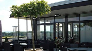 Un espacio al aire libre con mesas y sillas, un ventanal grande y un árbol.