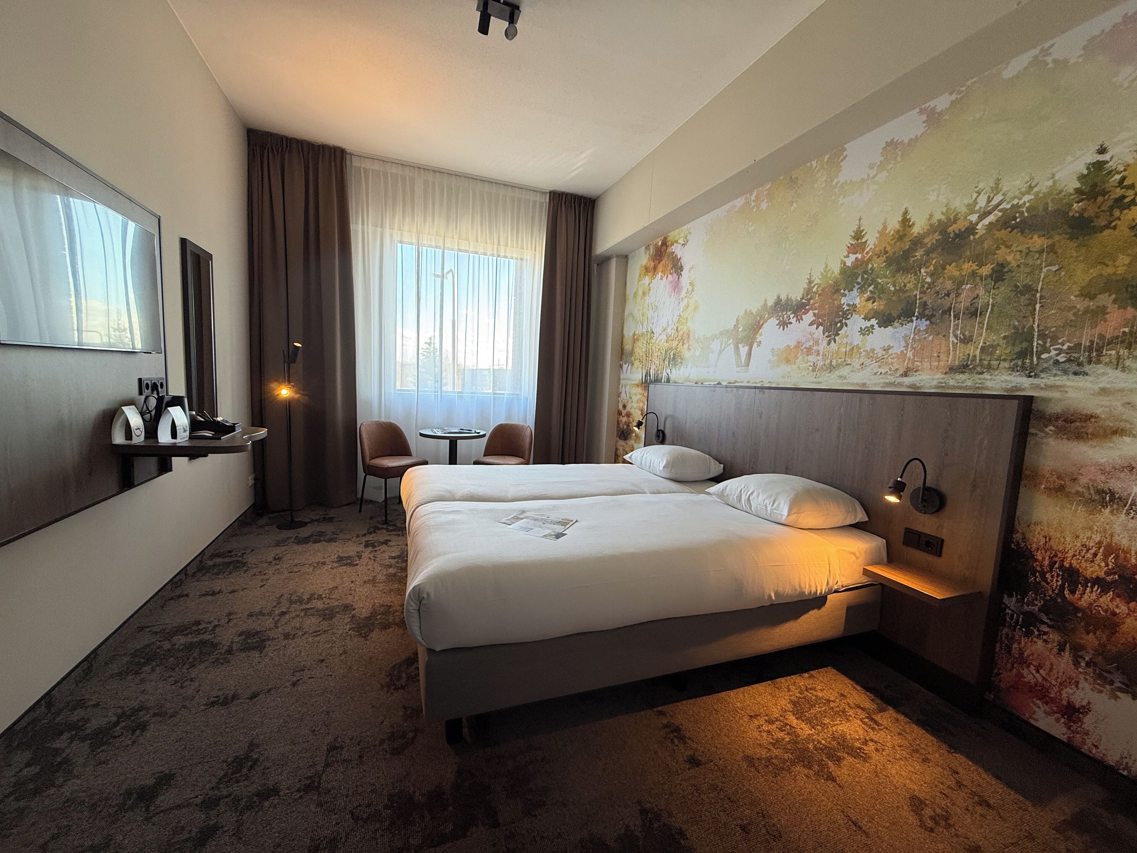 A hotel room with two beds, a desk with a chair, a small table, and a large window with curtains.