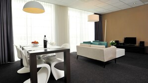 Una habitación de hotel moderna con zona de comedor, sofá, televisor y un ventanal con cortinas.