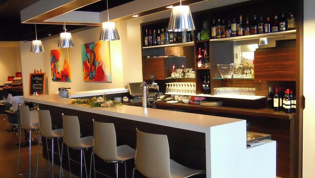 Un bar moderno con una barra alargada, varios taburetes, lámparas colgantes y estantes llenos de botellas.